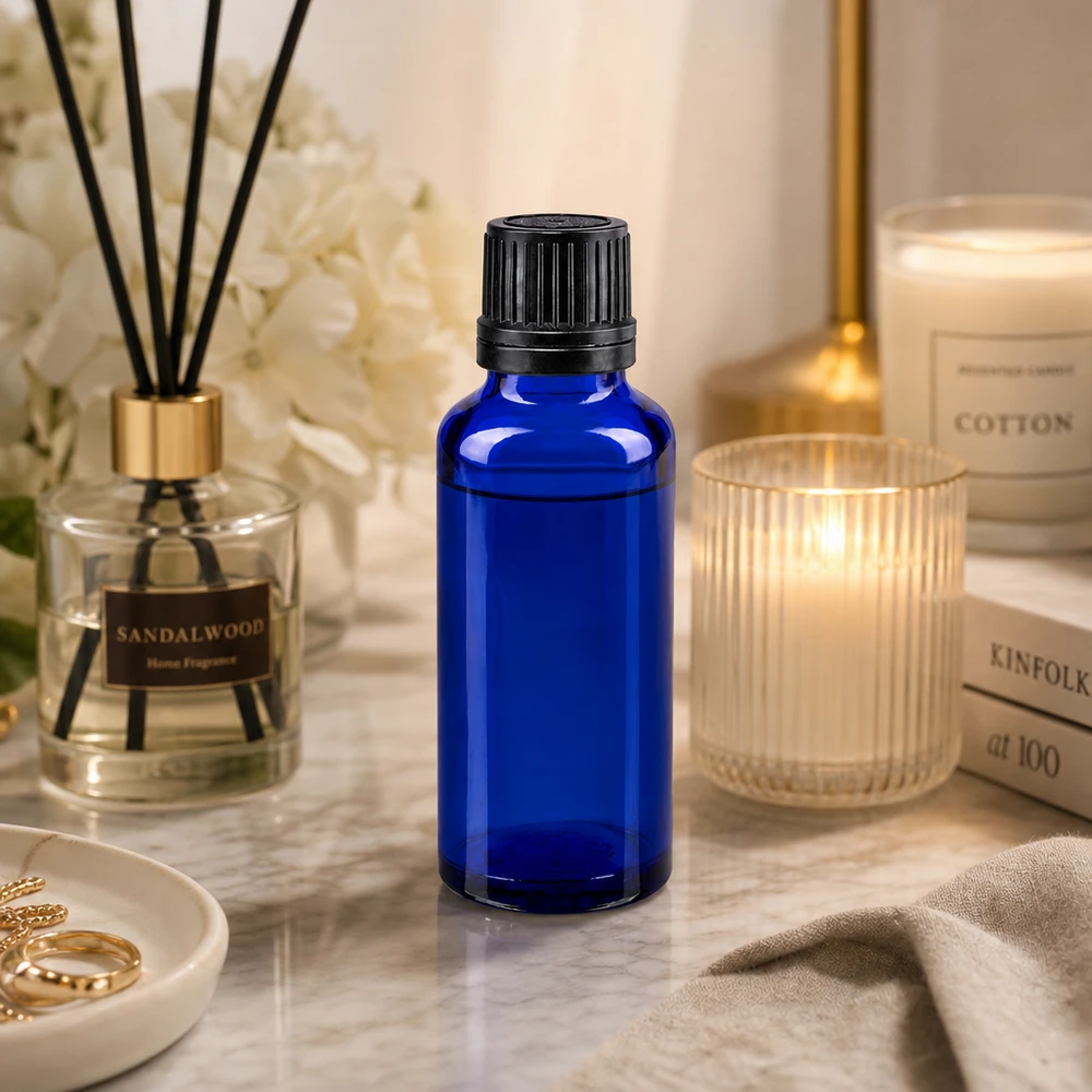 Frasco de vidro azul de 30 ml com tampa conta-gotas preta, em cima de uma superfície de mármore, ao lado de uma vela perfumada e de um difusor de aroma