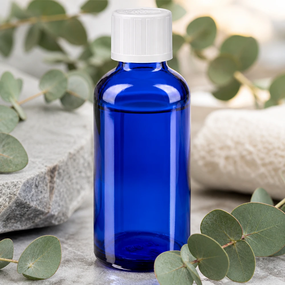 Frasco de farmácia azul cobalto de 50 ml com tampa KiSi branca, pousado sobre pedra natural entre ramos de eucalipto