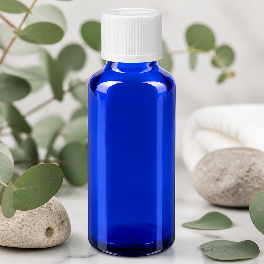 Frasco de farmácia azul cobalto de 30 ml com tampa KiSi branca sobre fundo de mármore, entre ramos de eucalipto