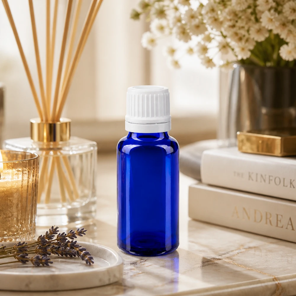 Frasco de vidro azul de 20 ml com tampa de encaixe branca sobre uma mesa de mármore, ao lado de um difusor de aroma para o ambiente