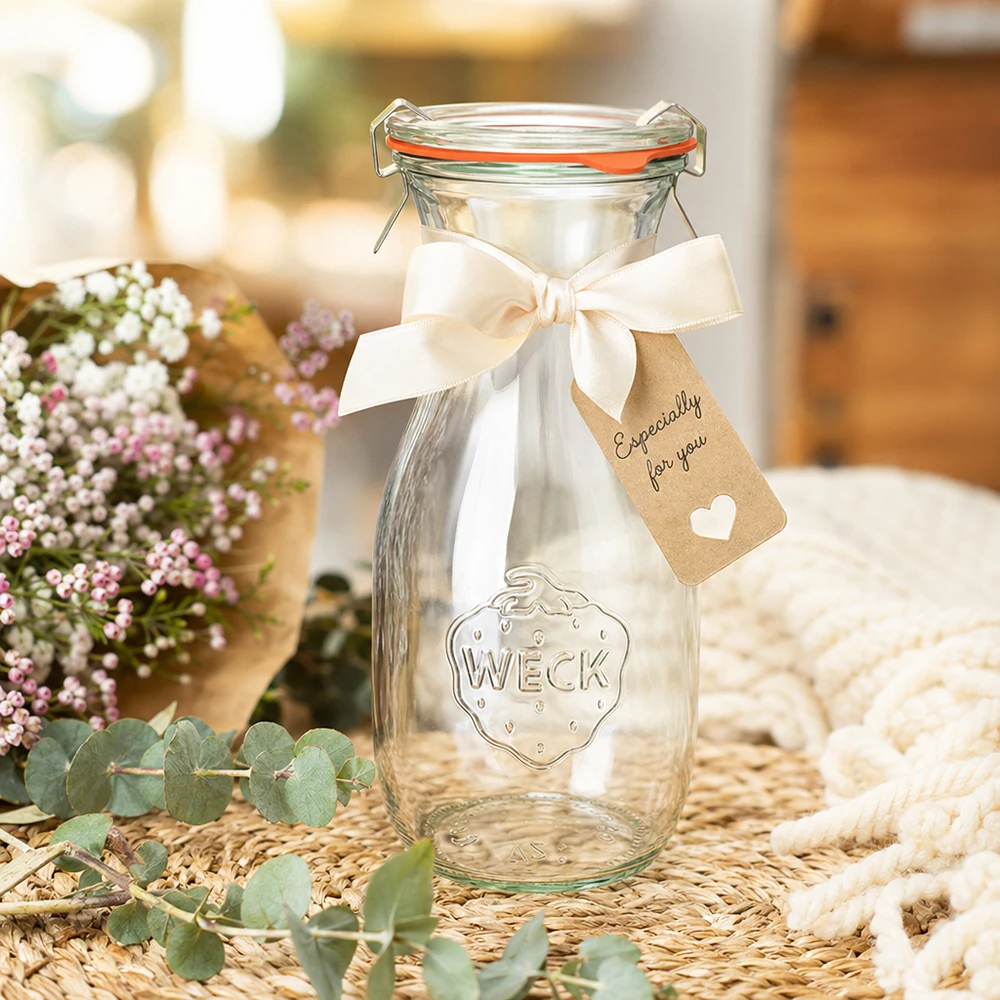 Garrafa de sumo WECK 530 ml RR60 decorada com um laço branco e uma etiqueta de oferta disposta entre flores
