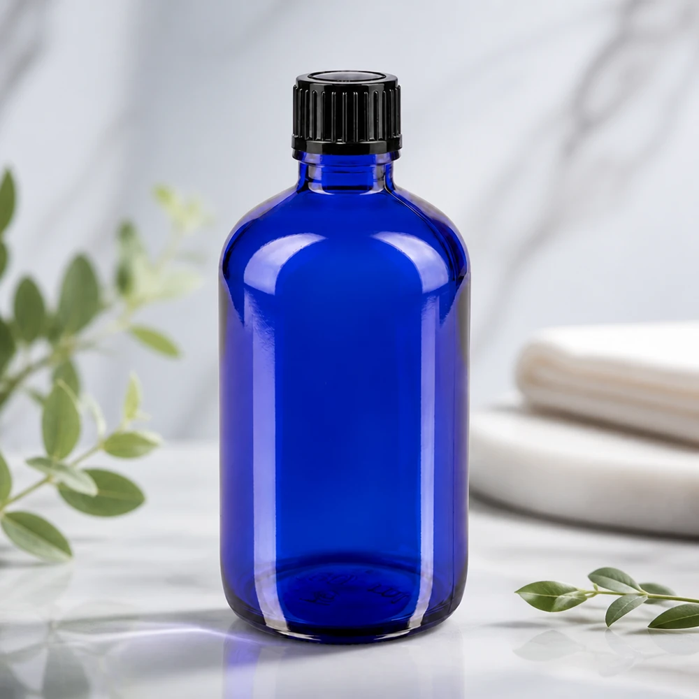 Frasco de farmácia azul 100ml com tampa de conta-gotas 1mm preto standard Garrafa de vidro azul de 100 ml com tampa preta estriada sobre uma superfície de mármore branco com ramos de eucalipto