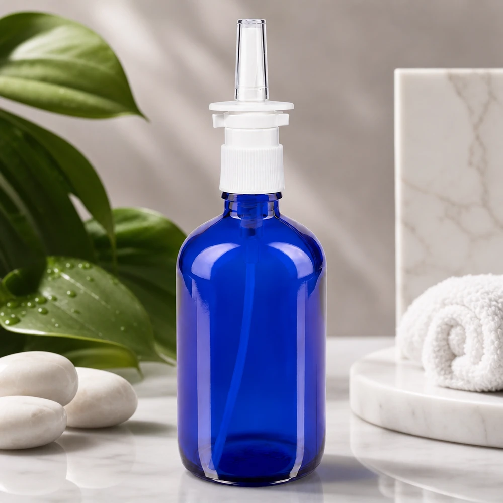 Frasco de farmácia azul 100ml atomizador nasal branco standard Frasco de vidro azul cobalto de 100 ml com bico pulverizador, sobre mármore branco, ao lado de pedras de bem-estar