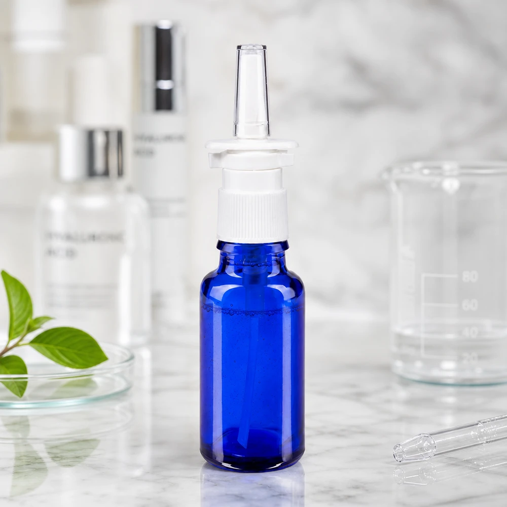 Frasco de spray nasal de 20 ml em vidro azul de farmácia com pulverizador branco, ao lado de um copo de vidro e de ácido hialurónico