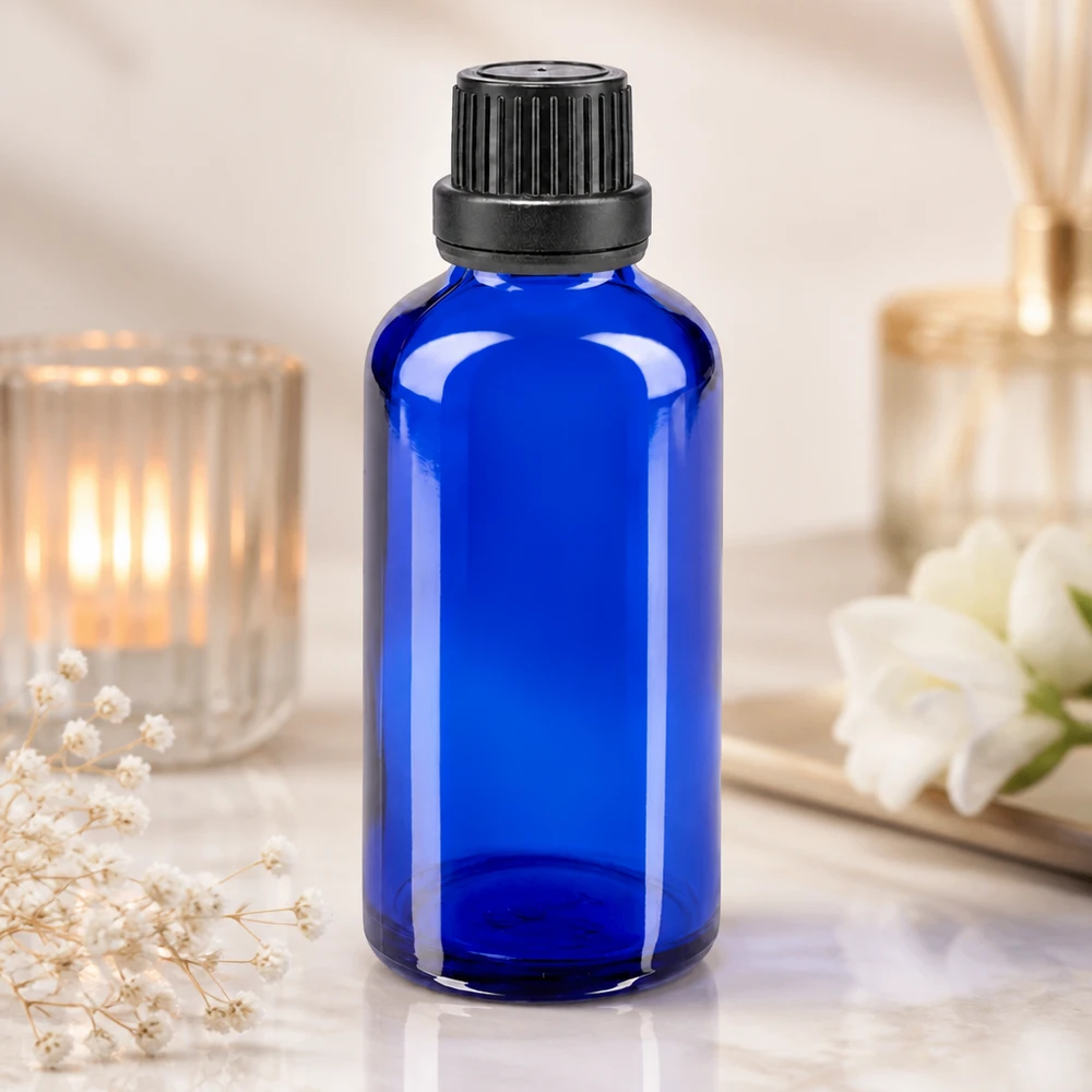 Frasco de vidro azul cobalto de 50 ml com tampa de rosca preta e conta-gotas, ao lado de uma vela e de flores