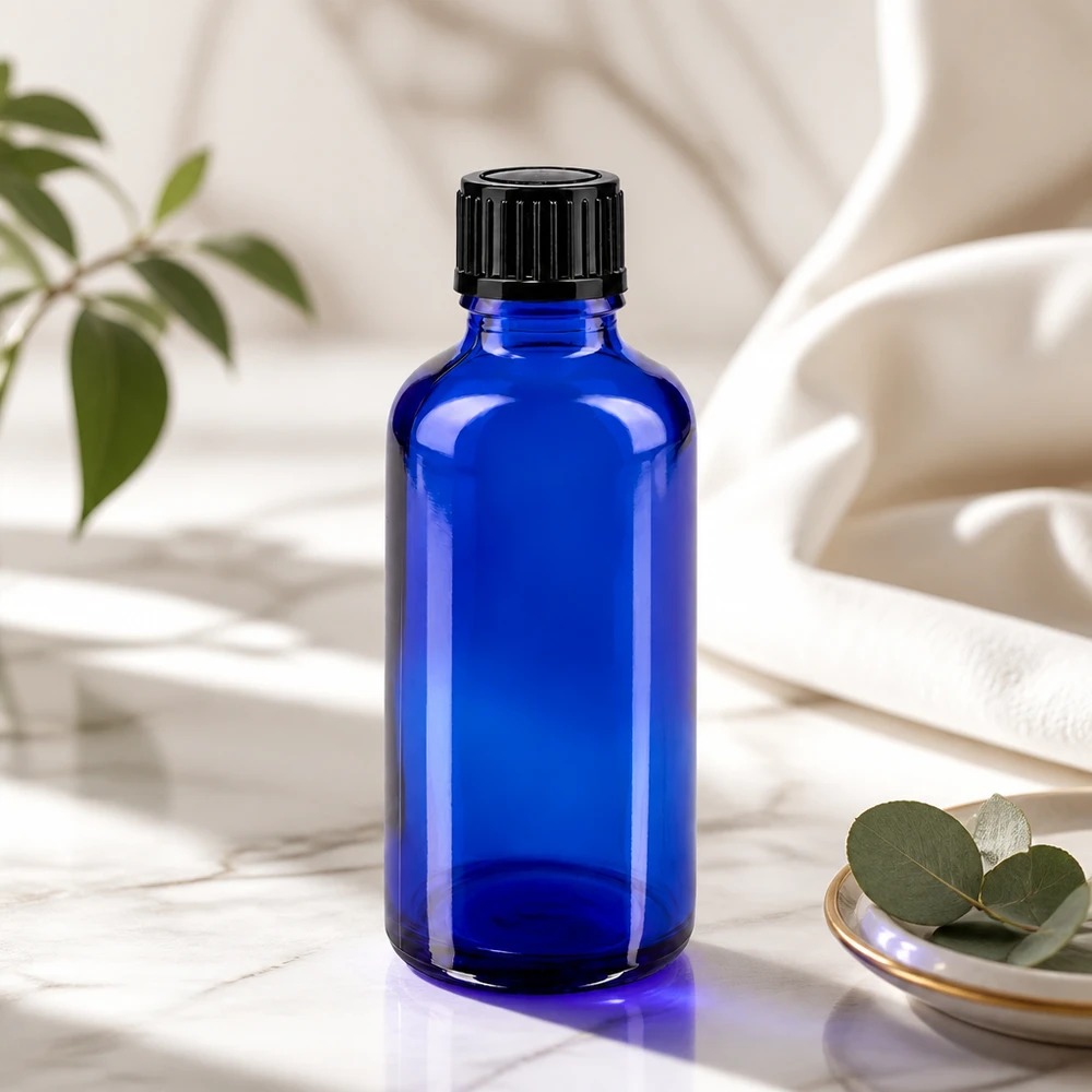 Garrafa de vidro azul de 50 ml com tampa preta sobre mármore, ao lado de folhas de eucalipto