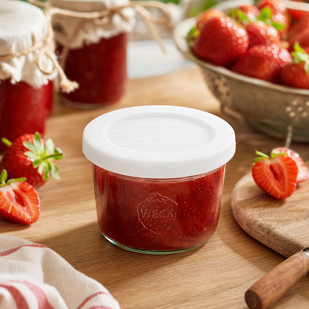 Copos WECK de 80 ml RR60 cheios de compota de morango numa tábua de madeira rodeada de morangos frescos e frascos de compota