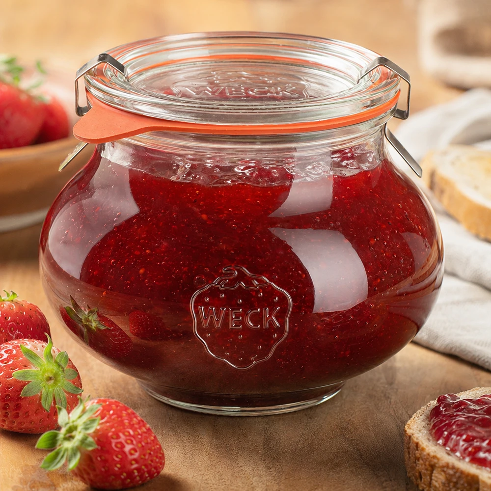 Frasco decorativo de 560 ml completo WECK RR80 Frasco decorativo WECK de 560 ml cheio de compota de morango caseira, tampa basculante fechada, rodeado de frutas e legumes frescos.