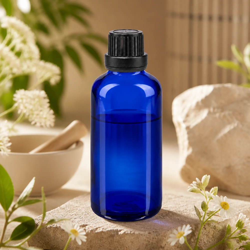 Frasco de vidro azul cobalto de 50 ml com tampa de rosca preta, apoiado sobre uma pedra natural com flores de camomila e um almofariz