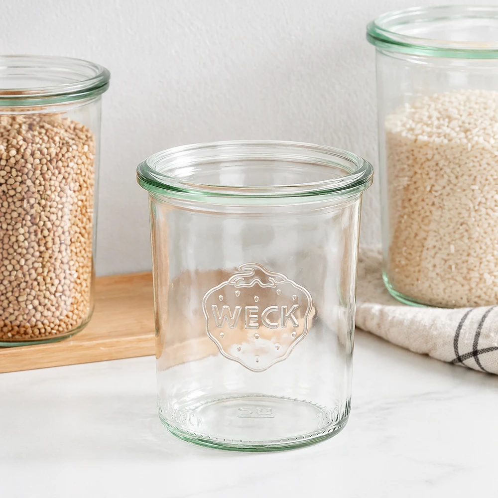 Copos de vidro de 160ml WECK RR60 Frasco de pedreiro WECK 160ml RR60 com rebordo verde e logótipo, frasco de pedreiro vazio ao lado de frascos de armazenamento cheios numa tábua de madeira