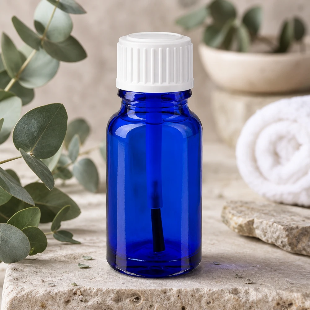 Frasco de vidro azul de 10 ml com um aplicador de pincel branco, ao lado de ramos de eucalipto sobre pedra natural