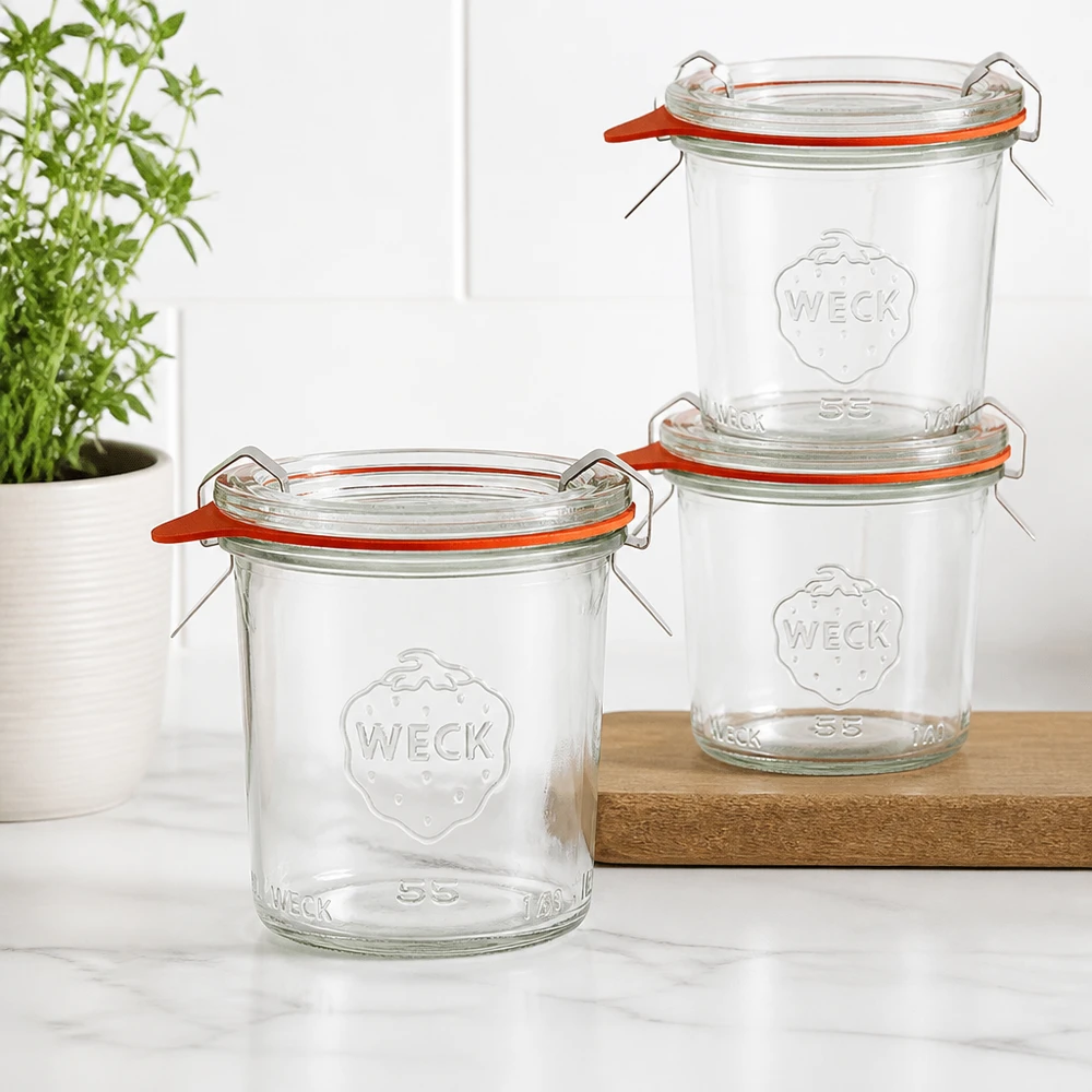 Copos de vidro de 140ml completos WECK RR60 Três copos WECK RR60 de 140 ml empilhados numa tábua de madeira numa cozinha moderna com uma planta verde em fundo