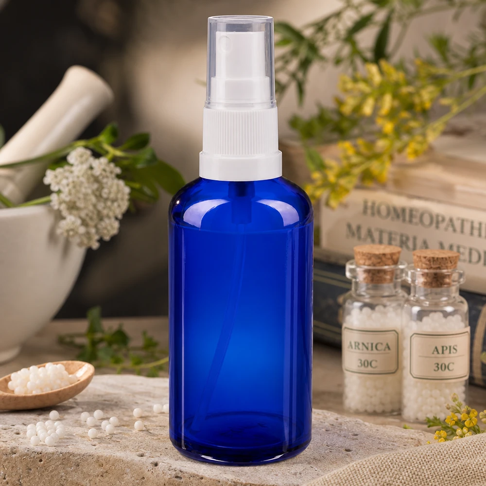 Frasco de vidro azul de farmácia com pulverizador de 50 ml e cabeça de pulverização branca, ao lado de glóbulos homeopáticos